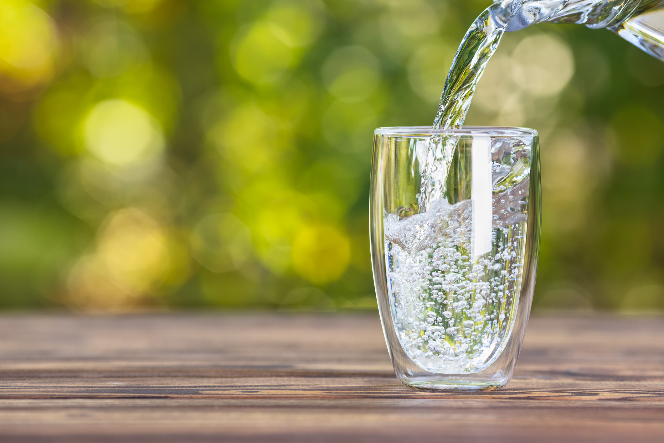 Water from a jug pouring into a glass on a wooden table outdoors.